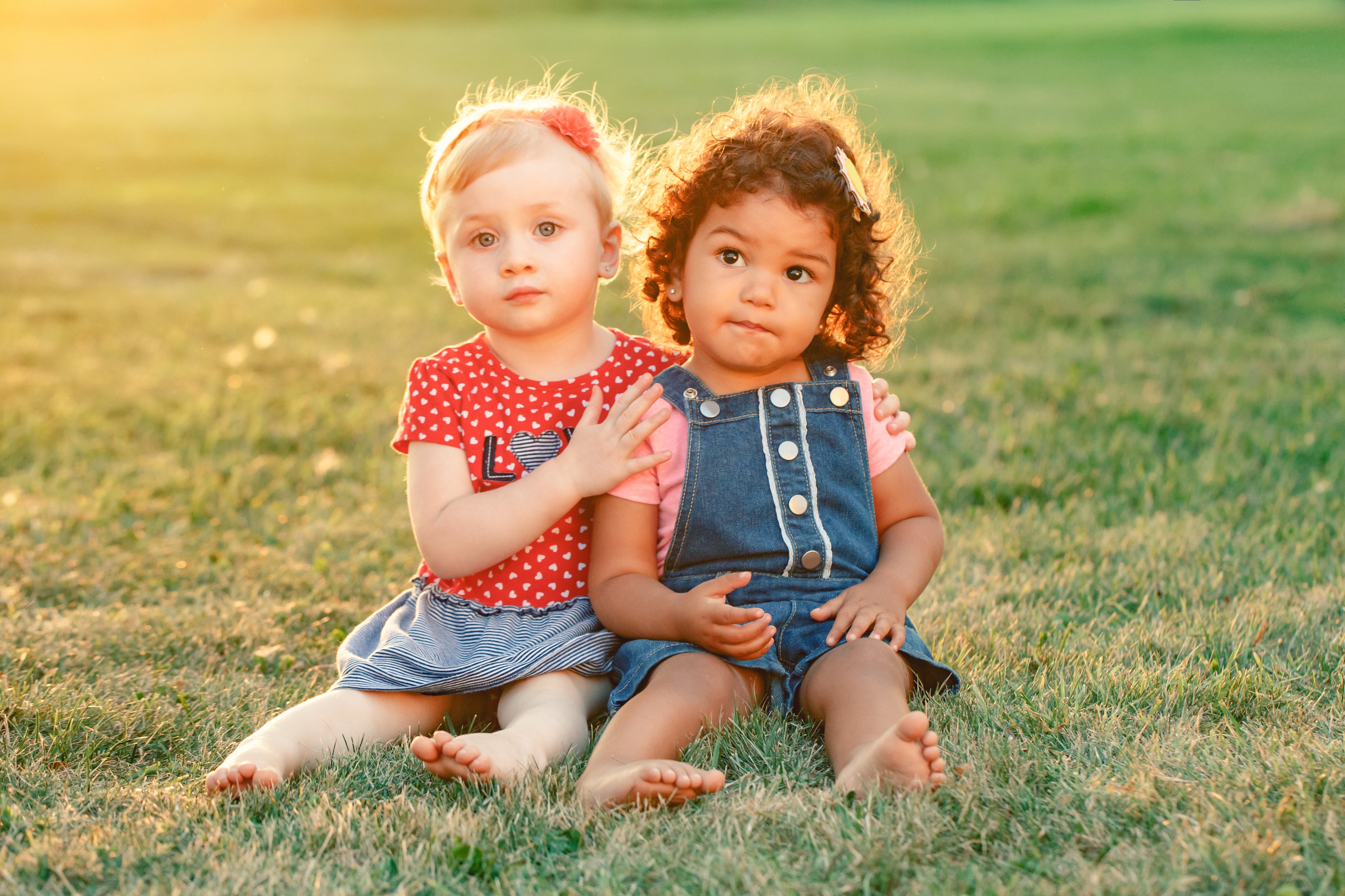 Two toddler girls sitting in the grass and embracing each other.