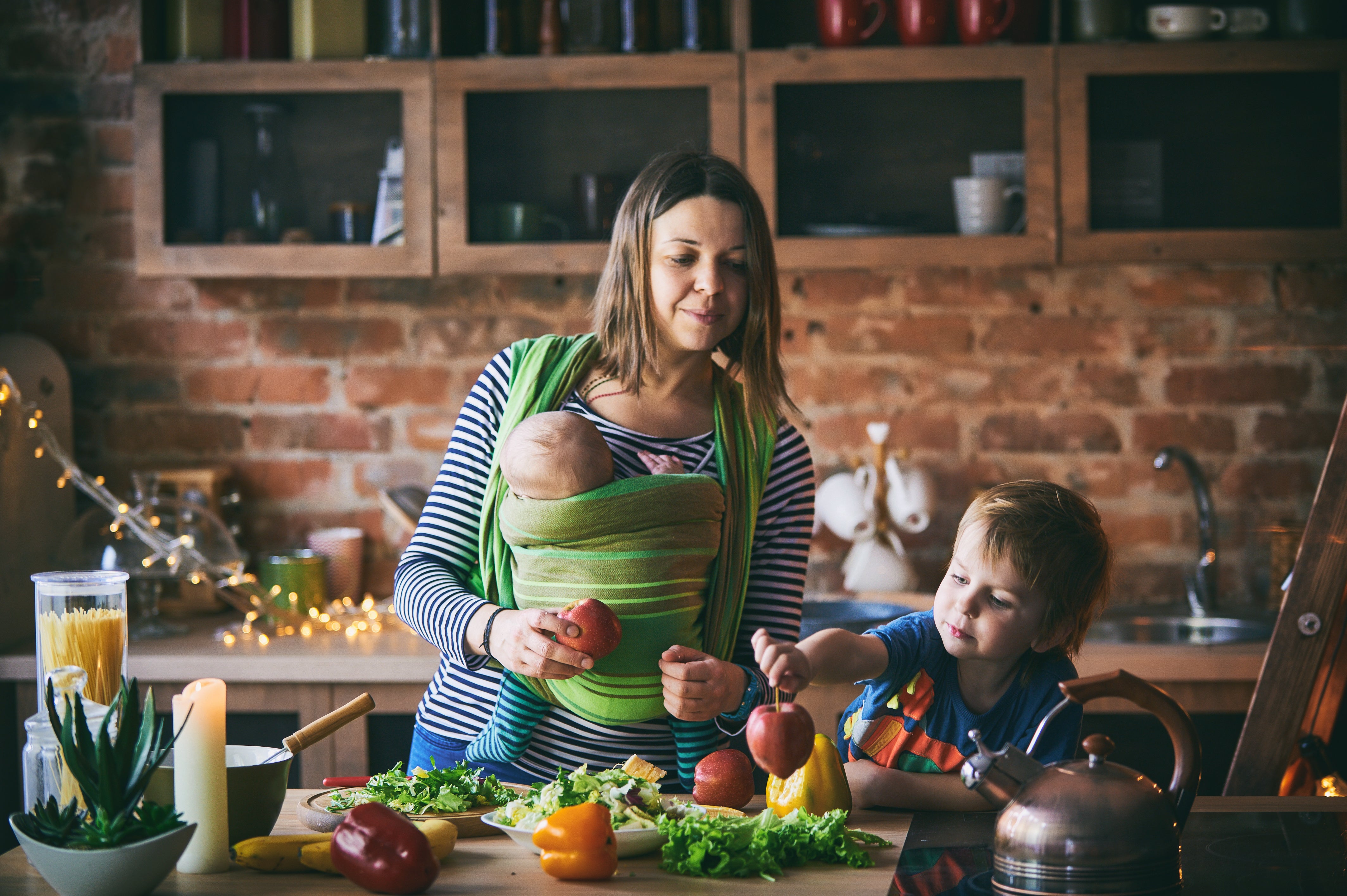 Mom baby-wearing an infant while cooking a healthy meal with toddler.