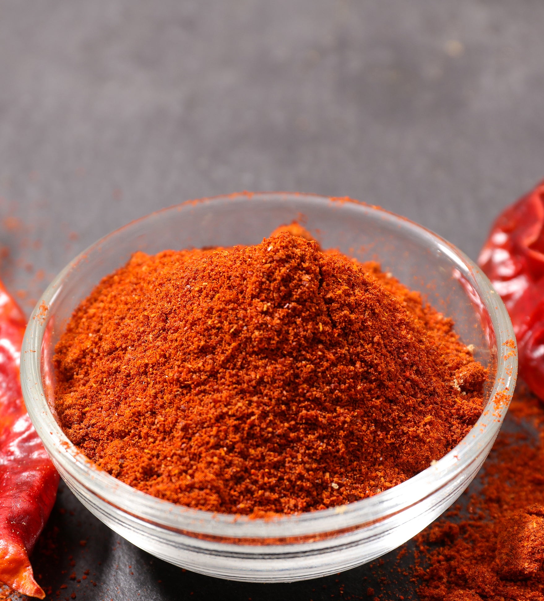 A close-up shot of a bowl of paprika powder, and vibrant red paprika peppers against a grey background with a focus on its textured surface and stem.