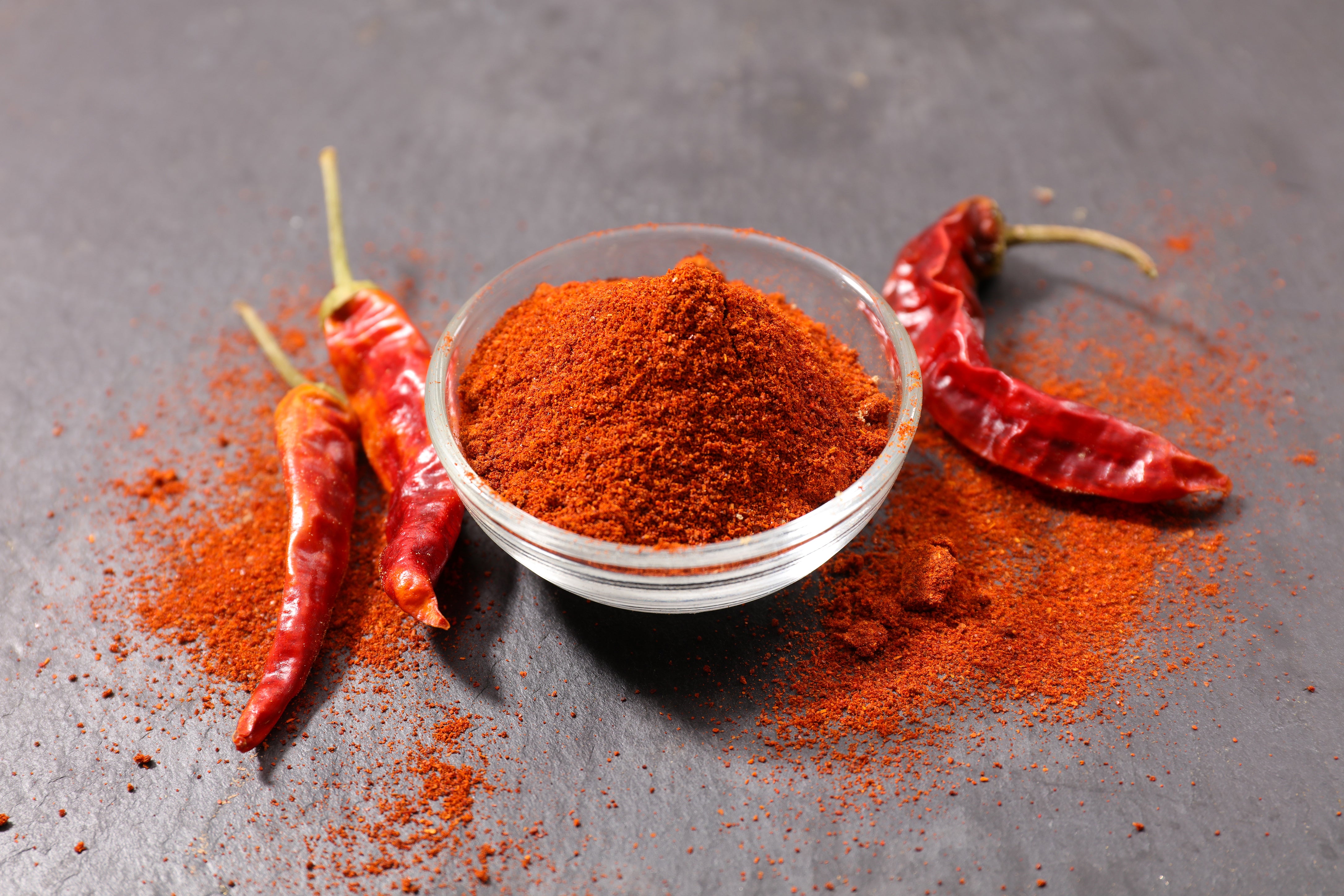 A close-up shot of a bowl of paprika powder, and vibrant red paprika peppers against a grey background with a focus on its textured surface and stem.