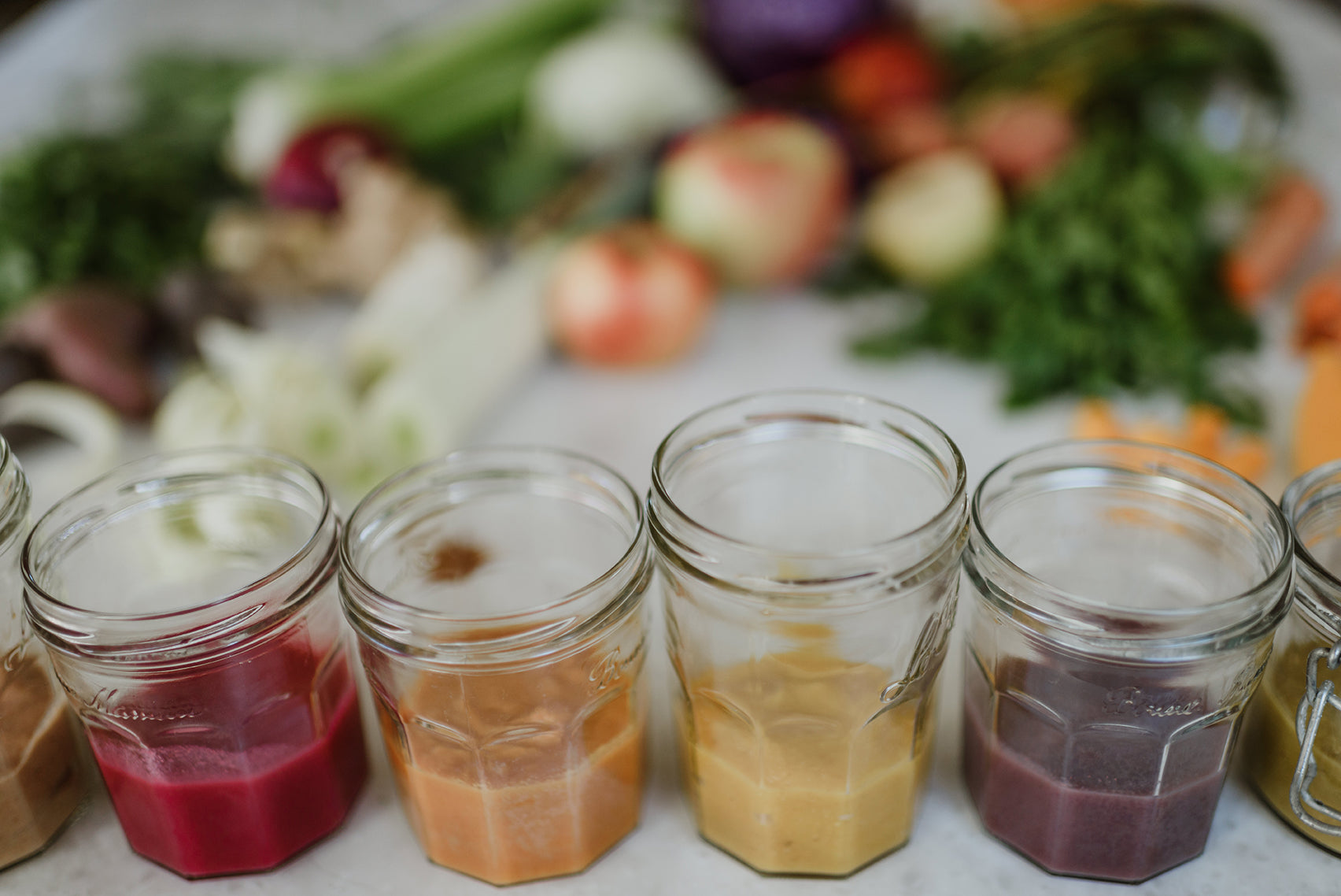 An assortment of baby food jars, each with different vibrant colors indicating various fruit and vegetable purees, arranged in a semi-circle on a white background.